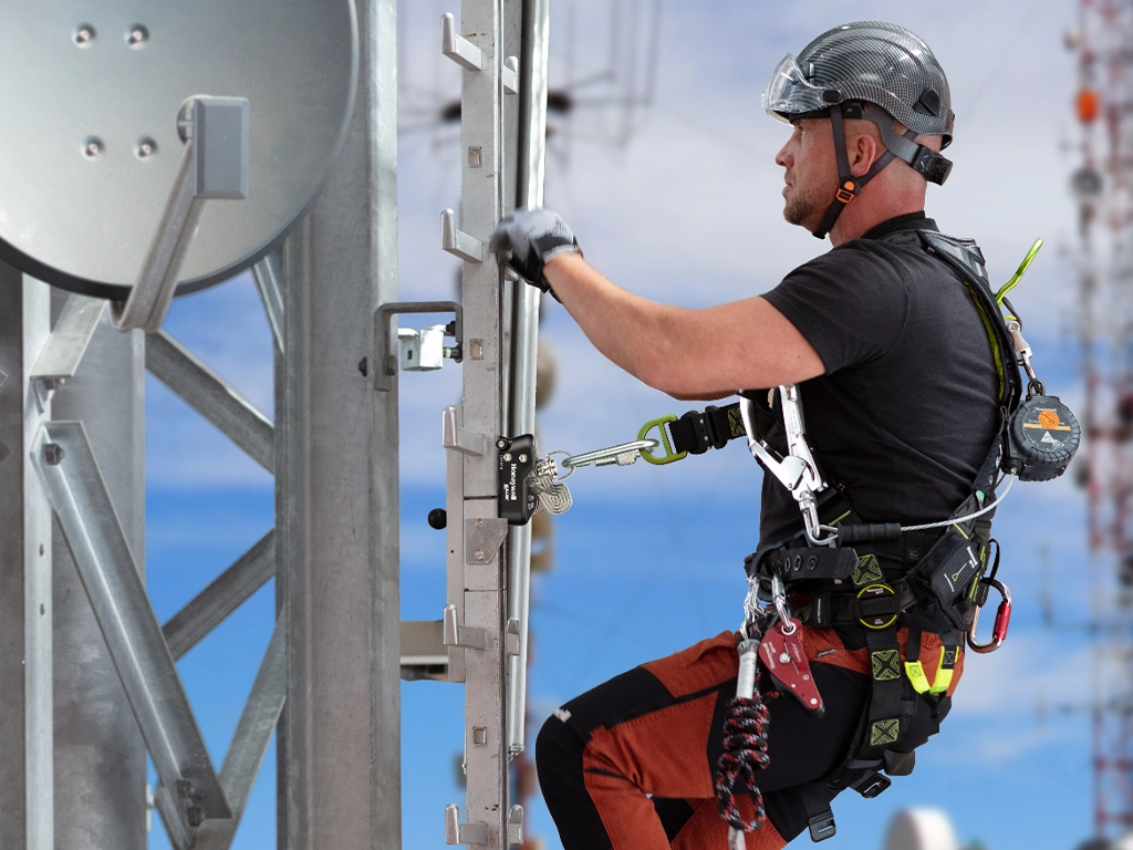 Ein Industriekletterer in voller Sicherheitsausrüstung steigt an einer metallischen Leiter auf einen Telekommunikationsmast. Er trägt einen schwarzen Schutzhelm mit Visier, ein schwarzes T-Shirt, eine rot-schwarze Arbeitshose und Schutzhandschuhe. Er absolviert eine Schulung zur persönlichen Schutzausrüstung gegen Absturz